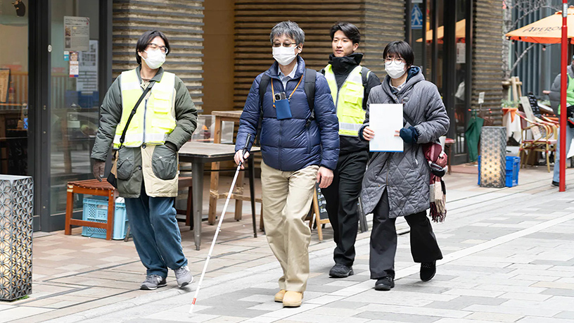 街の中の歩道を、白杖を持つ人を含む複数人が並んで歩いている様子